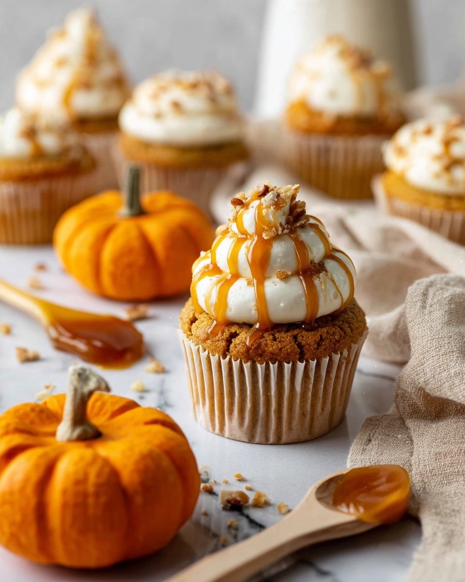 A close-up view of a single pumpkin cupcake resting on a white marbled surface, with a golden brown base wrapped in a white cupcake liner. On top, three tall swirls of creamy white frosting create a soft, smooth texture, drizzled with an amber caramel sauce that glistens. Small pecan crumbs are sprinkled lightly over the caramel. Surrounding the cupcake are two small orange pumpkins with rugged stems, a beige cloth to the right, a wooden spoon with caramel next to the cupcake, and a few other cupcakes blurred in the background. The overall color scheme mixes warm tones of orange, caramel, and white, with a cozy fall feel. Photo taken with an iphone --ar 4:5 --v 7