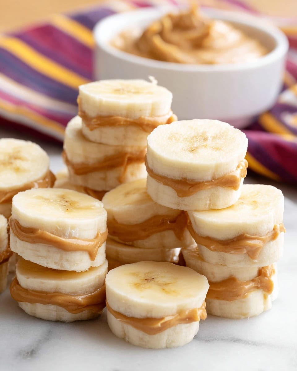 A close-up view of multiple small stacks of peeled banana slices layered with creamy peanut butter in between each slice, forming mini sandwich-like pieces. The banana slices are pale yellow with a soft texture, while the peanut butter layers are smooth and light brown, slightly oozing out from the edges. The stacks are arranged loosely on a clean white marbled surface. In the background, a white bowl filled with peanut butter sits on a striped cloth with red, purple, and yellow lines, slightly blurred to keep focus on the banana and peanut butter stacks in the foreground. photo taken with an iphone --ar 4:5 --v 7