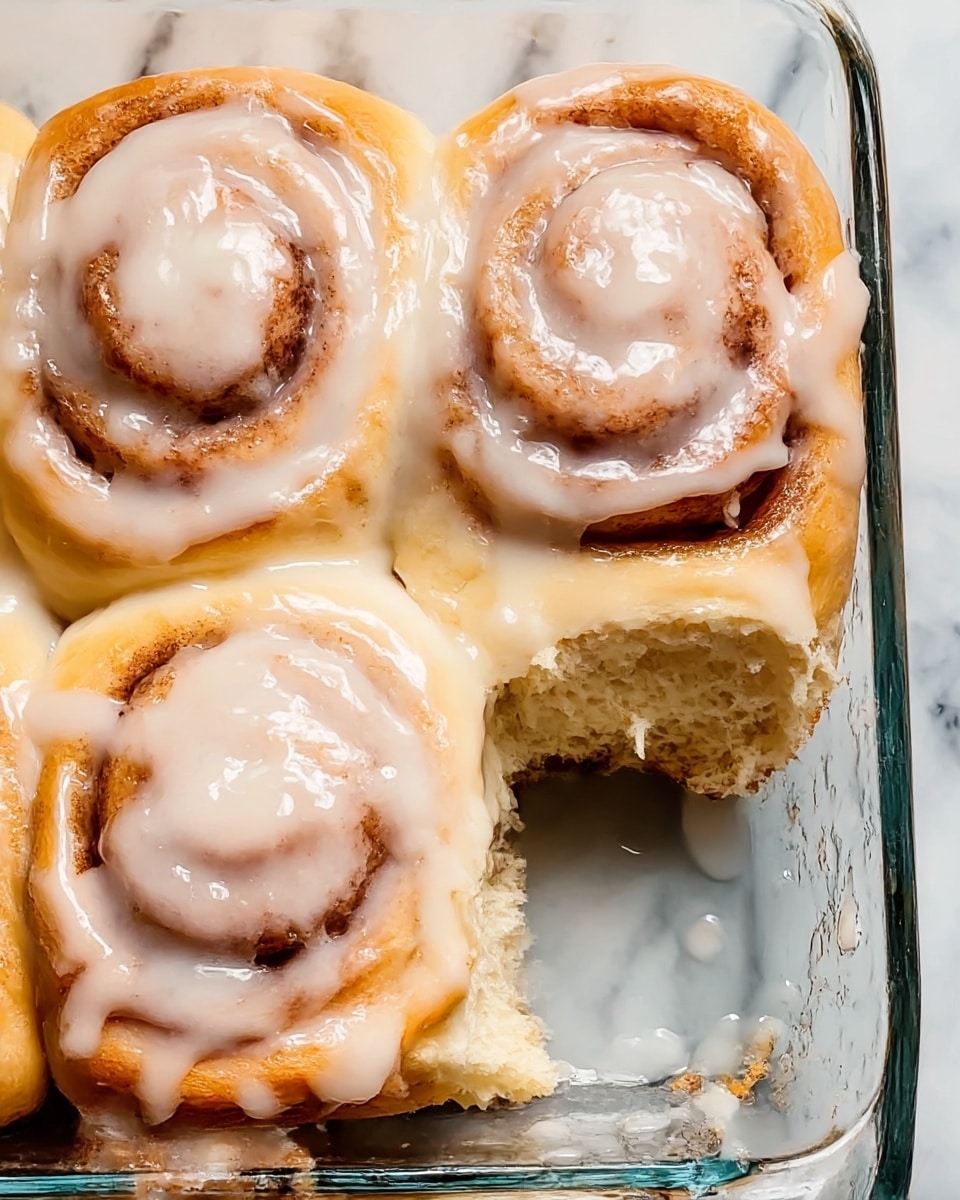 Four cinnamon rolls with a thick layer of white glaze cover the top, each roll showing a soft golden brown spiral pattern under the shiny icing. The rolls are close together in a clear glass baking dish, with one roll partly eaten showing a fluffy, light beige inside. The glaze drips slightly down the sides of the rolls. The baking dish sits on a white marbled surface. photo taken with an iphone --ar 4:5 --v 7
