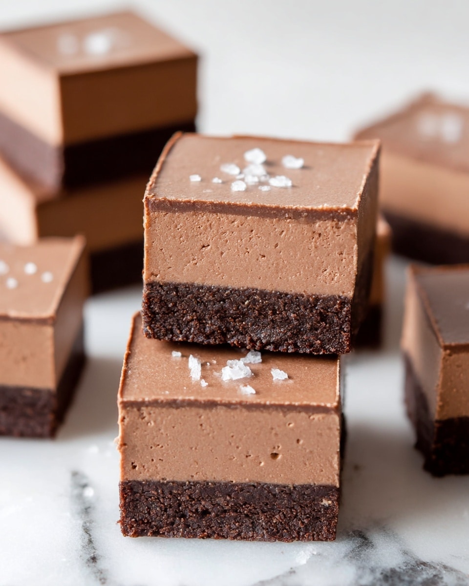 The image shows a close-up of several square dessert pieces stacked and scattered on a white marbled surface. Each piece has two layers: a smooth, thick top layer of light brown mousse-like chocolate, and a firm, crumbly dark chocolate base layer. The top surfaces have a few small salt flakes sprinkled lightly. The stacked squares are sharp-edged and cleanly cut, highlighting the thickness and texture of each layer. Photo taken with an iphone --ar 4:5 --v 7